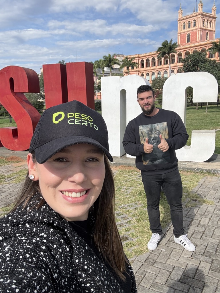 Pâmela de boné em primeiro plano e Samuel ao fundo fazendo sinal de positivo, com o Palacio de los López atrás, em Asunción
