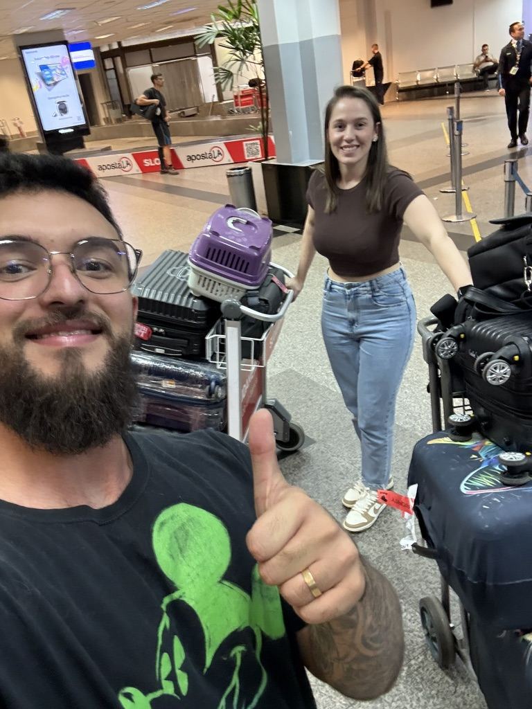 Ele sorrindo com sinal de positivo no aeroporto, ela ao fundo com as malas