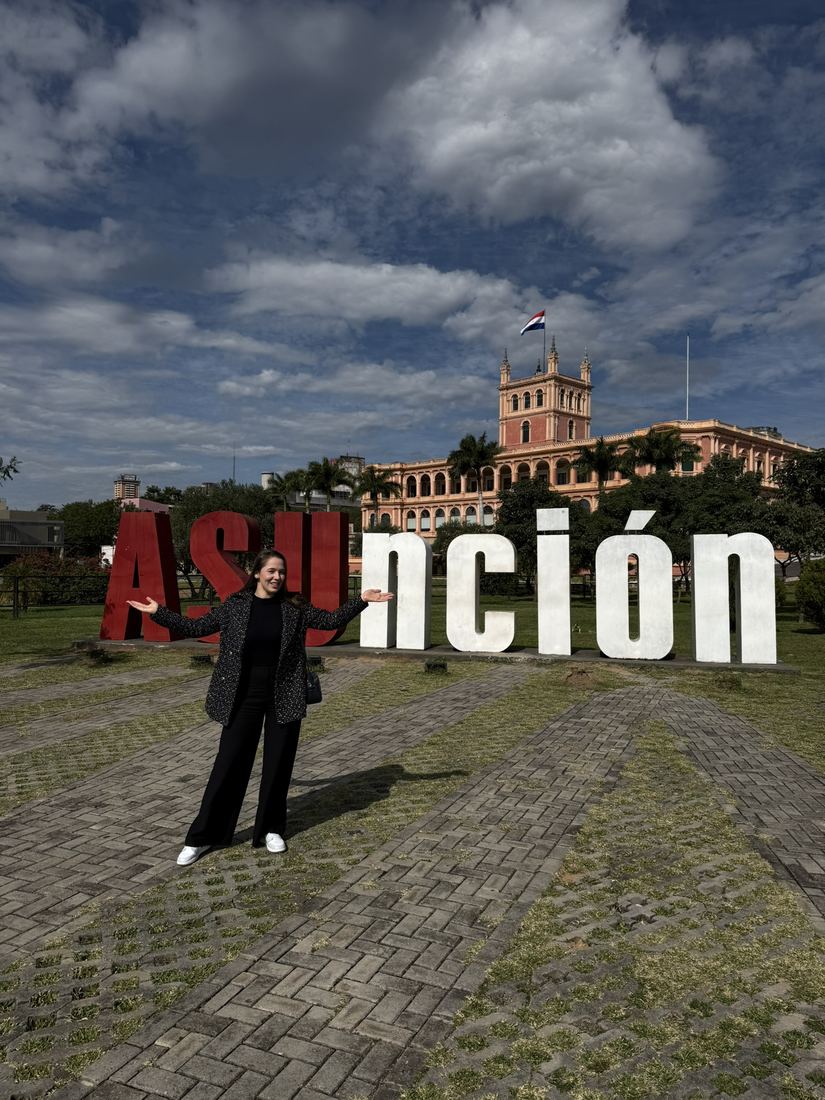 Ela em pé em frente ao letreiro de Asunción, com um palácio ao fundo