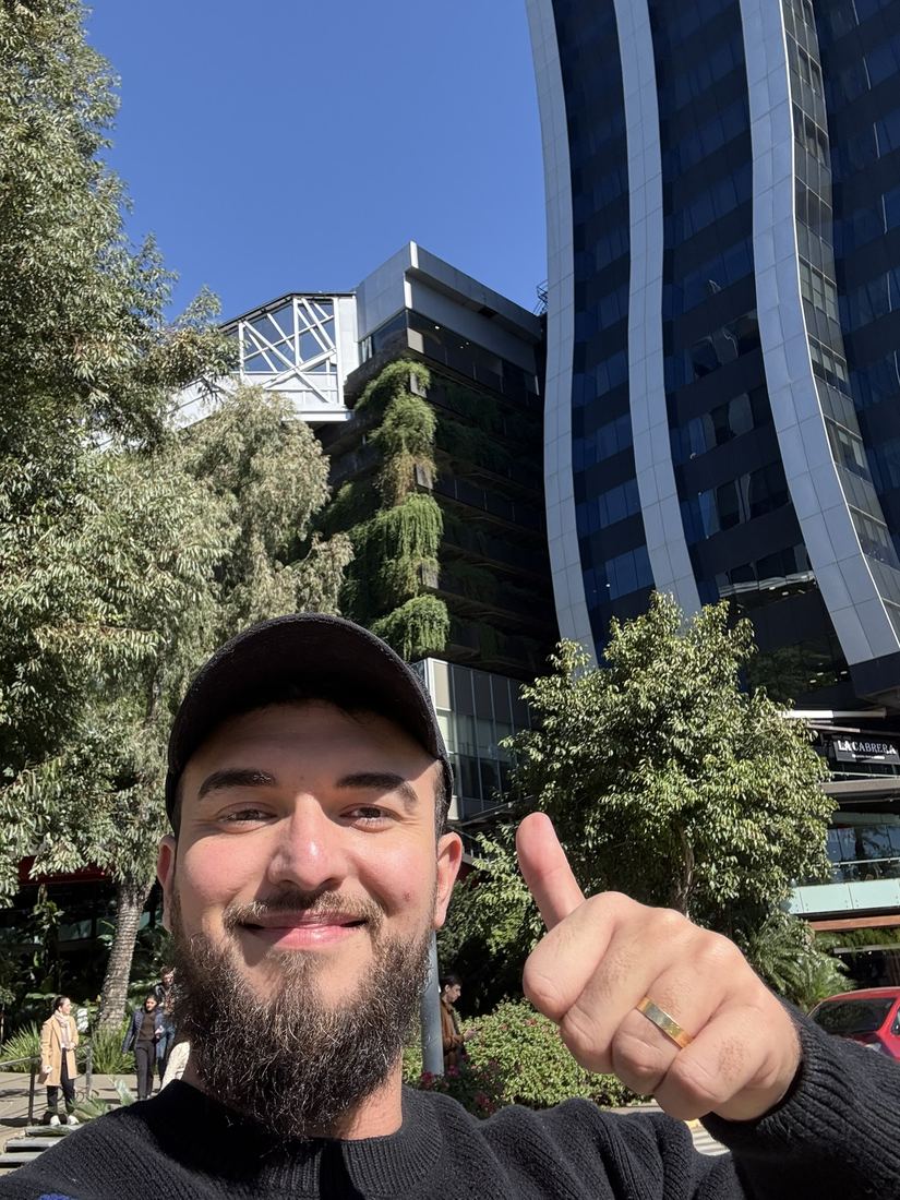Samuel fazendo sinal de positivo em frente a prédios espelhados e árvores