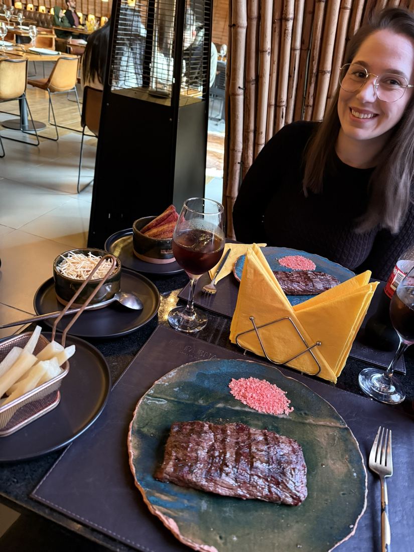 Ela sorrindo à mesa de um restaurante com um prato de carne